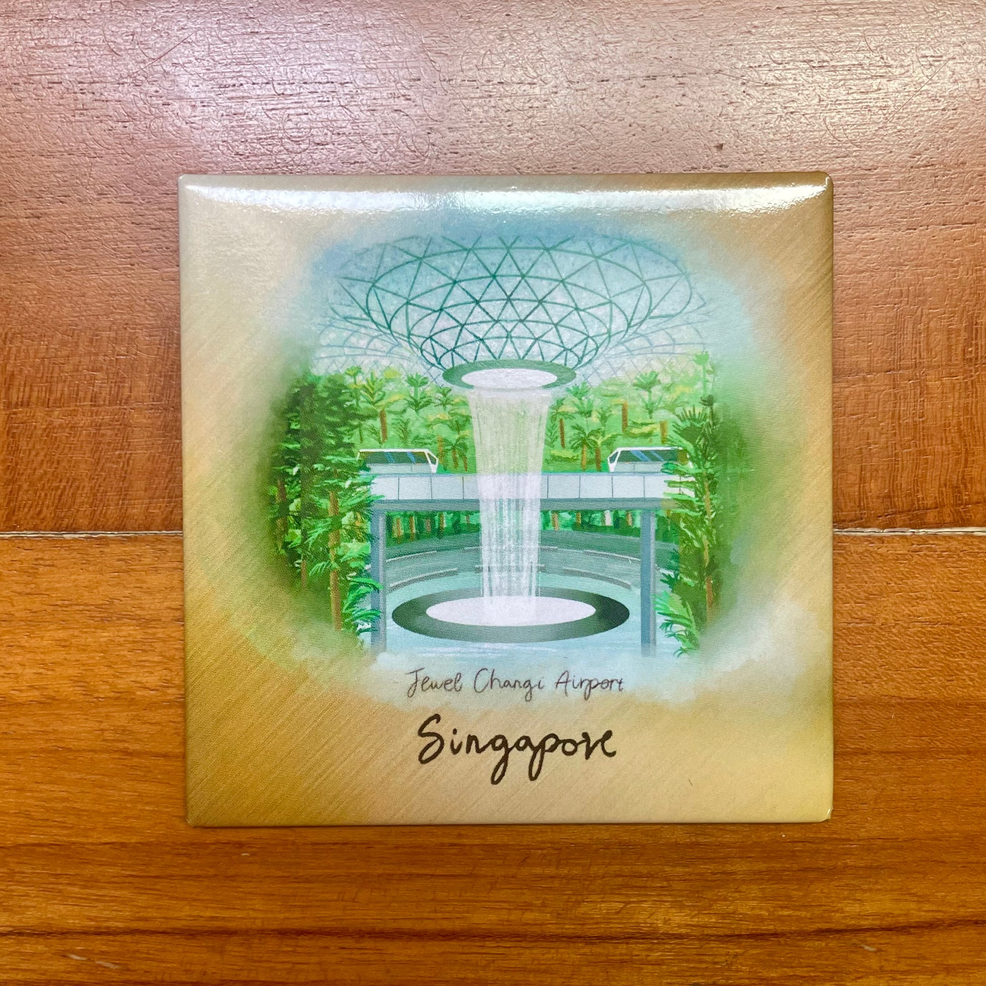 A round magnet featuring the Jewel Changi Airport with a depiction of a rain vortex in the center, surrounded by greenery and the airport's name 'Singapore' at the bottom.