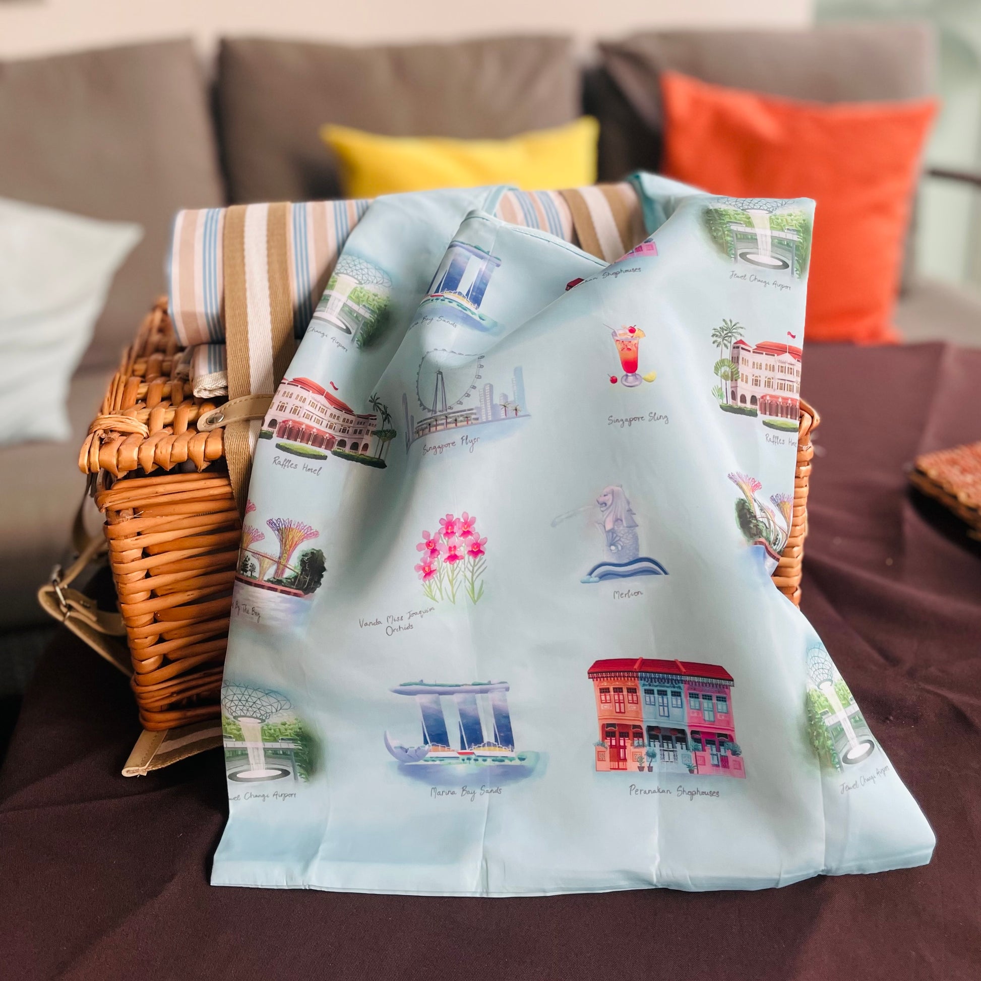 A foldable shopping tote with a design featuring various iconic landmarks and scenes in Singapore, placed over a picnic basket on a table.