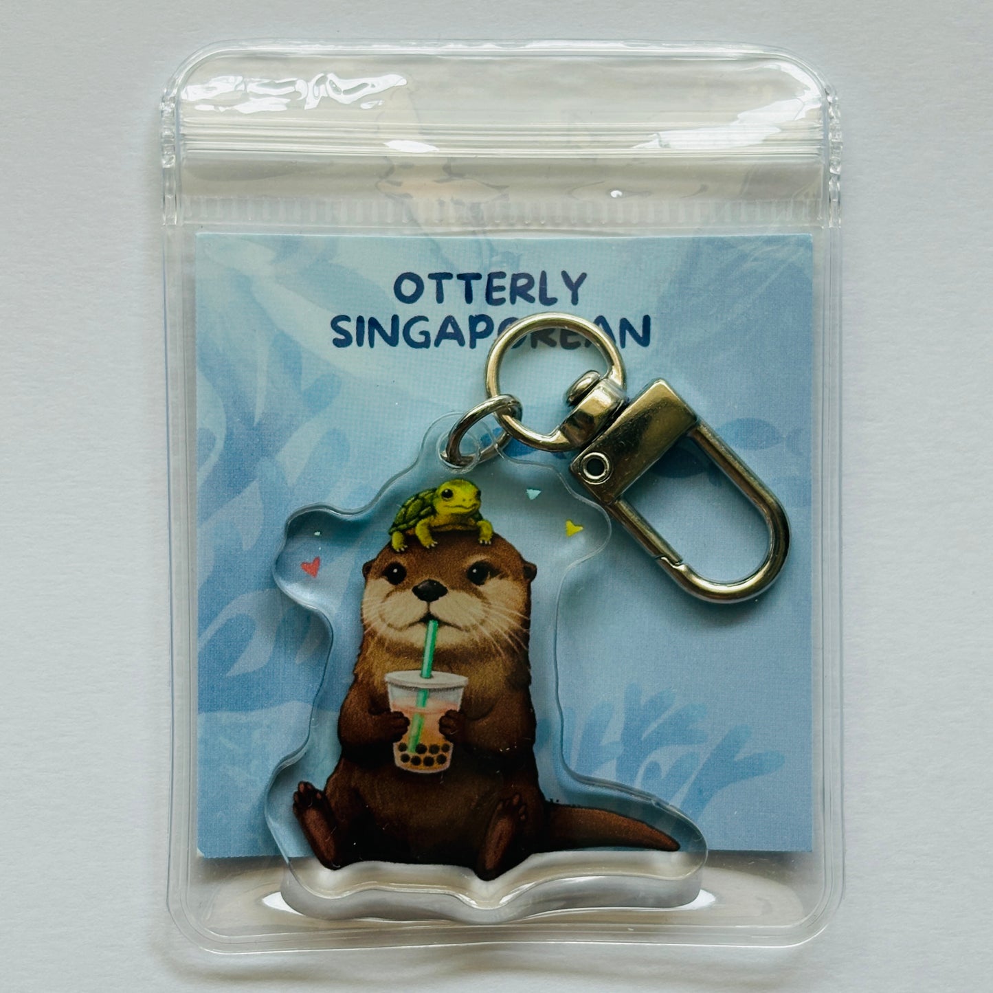 Keychain with otter design holding a bubbletea drink, branded 'Otterly Singaporean', on a blue card.