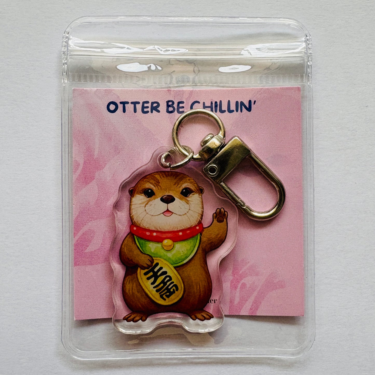 Keychain with otter and fortune coin design on a pink card labeled 'Otter Be Chillin''.