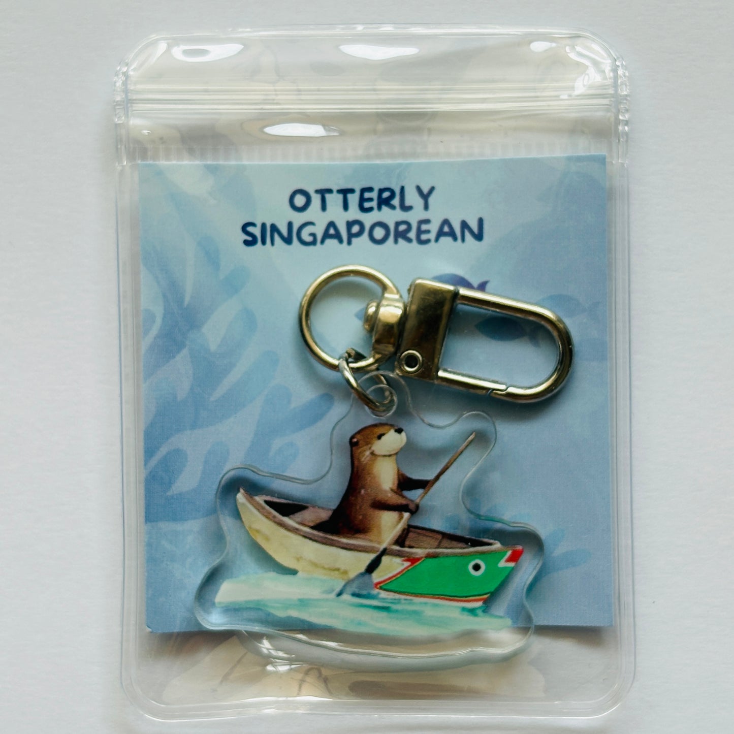 Keychain with an otter in a boat on a card labeled 'Otterly Singaporean'.